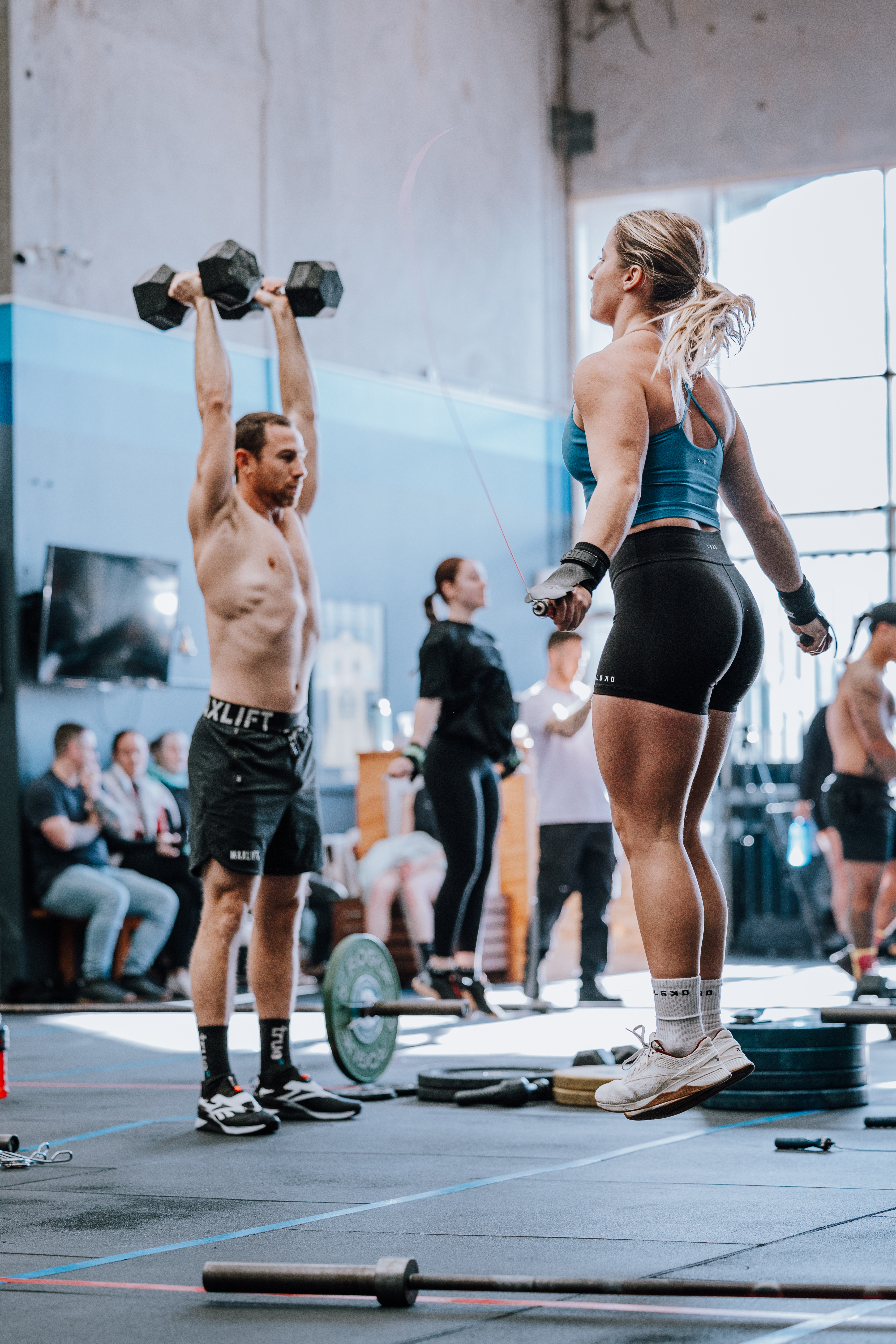 A fitness gym interior featuring two individuals engaged in a workout; one man is lifting dumbbells while a woman performs jump rope exercises.